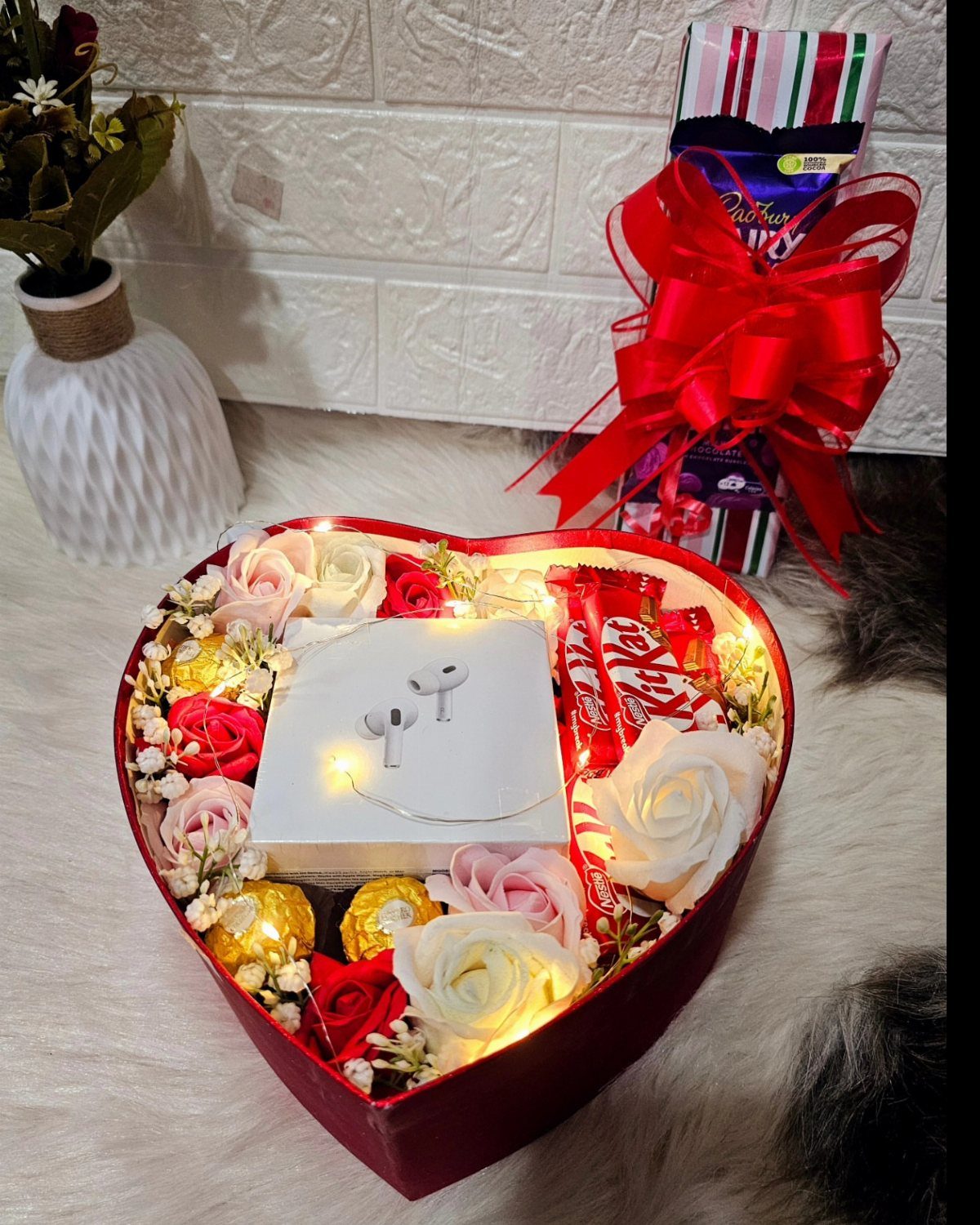 Heart-shaped red gift box with roses, chocolates, and wireless earbuds on white fur.