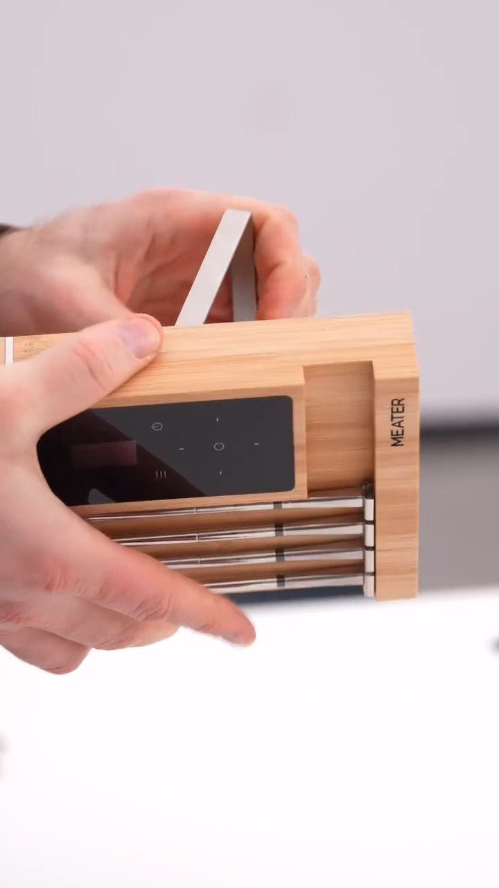 Hands holding a wooden MEATER smart meat thermometer charging base with metallic probes.