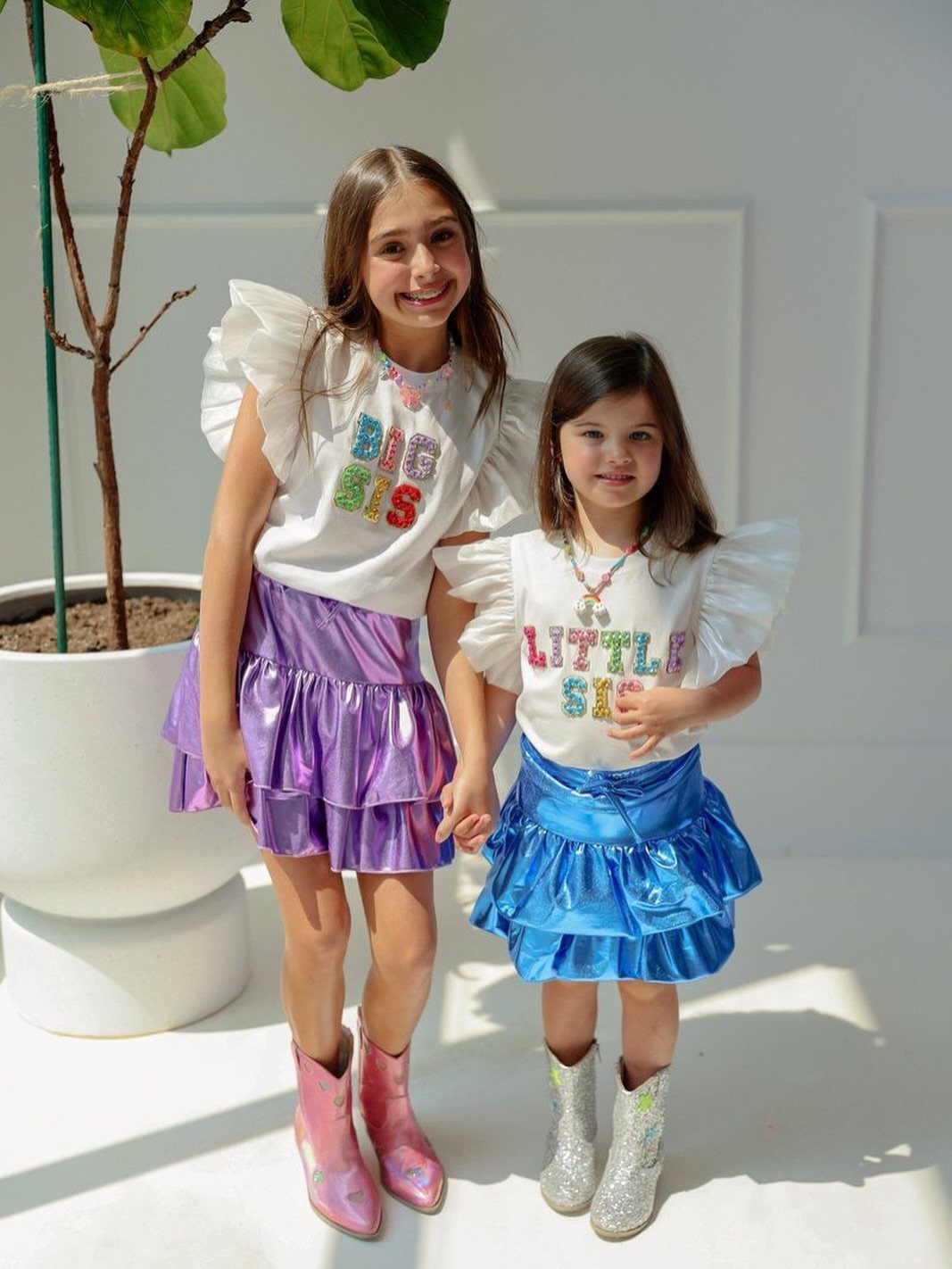 Two sisters wearing matching white ruffled shirts and metallic tiered skirts holding hands.