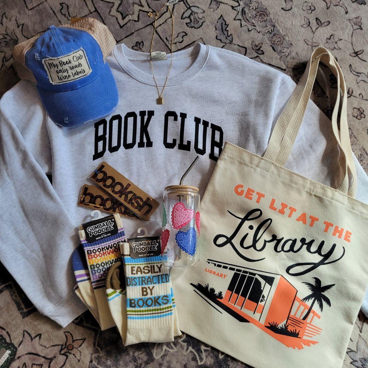 White Book Club sweatshirt surrounded by a tote bag, cap, and bookish accessories.