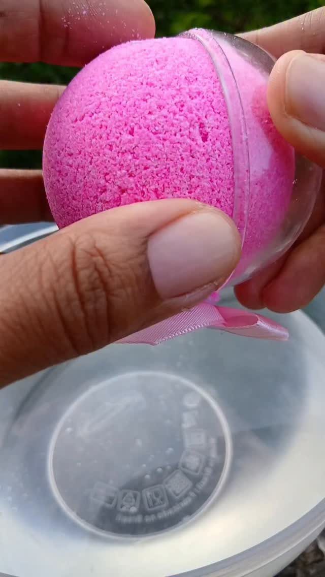 Bright pink spherical bath bomb in a clear plastic shell held by hands.