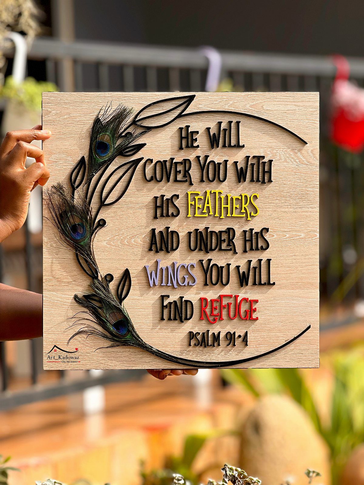 Wooden plaque with Psalm 91:4 and peacock feather art held by a hand