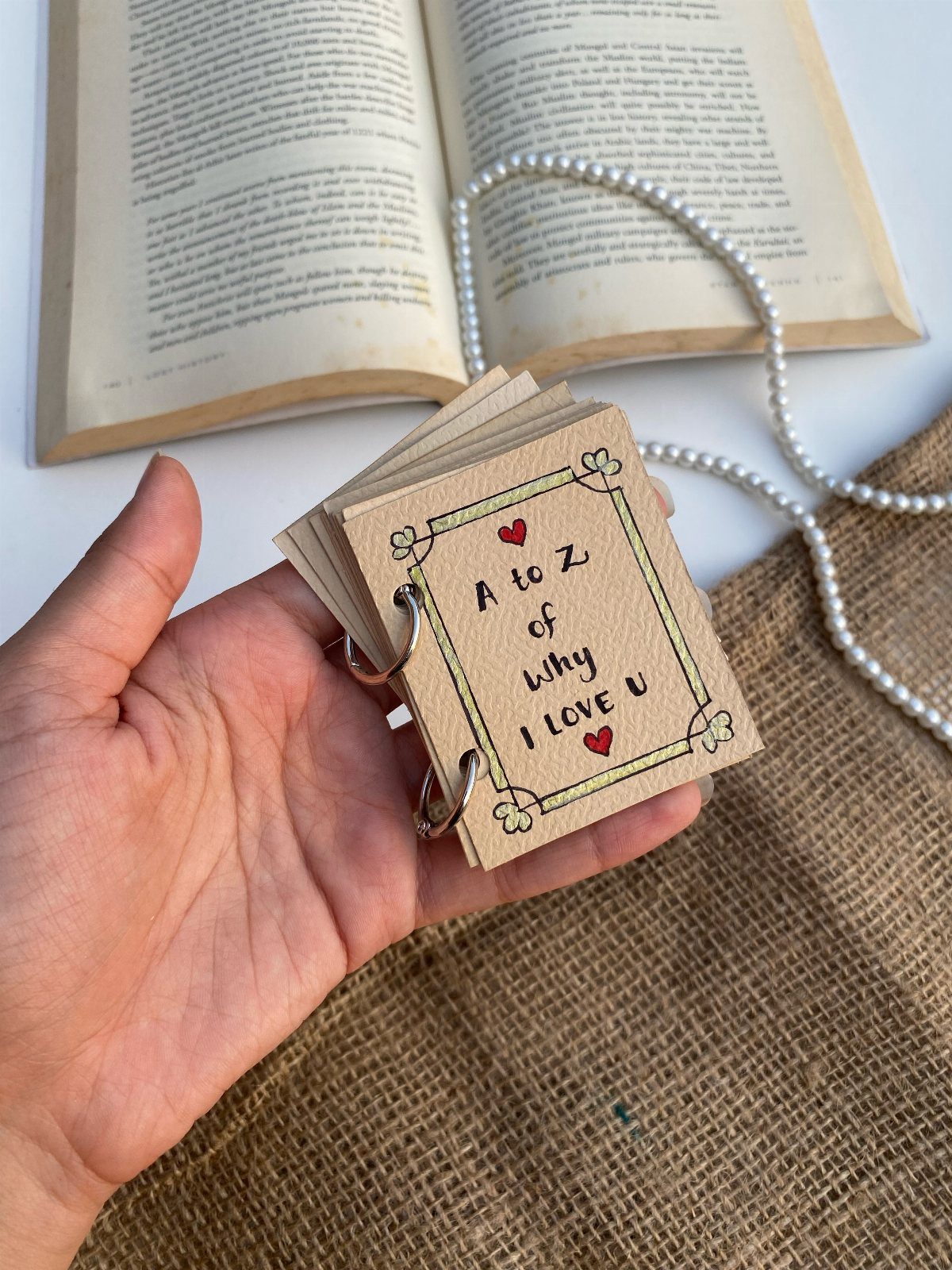 Handmade ring-bound love booklet held over an open book and pearl necklace.