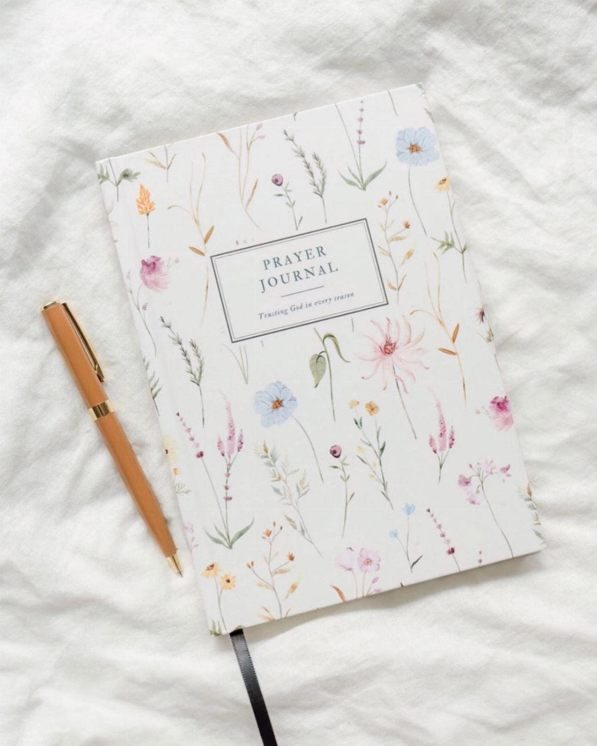 Floral prayer journal and gold pen resting on a white textured fabric background.