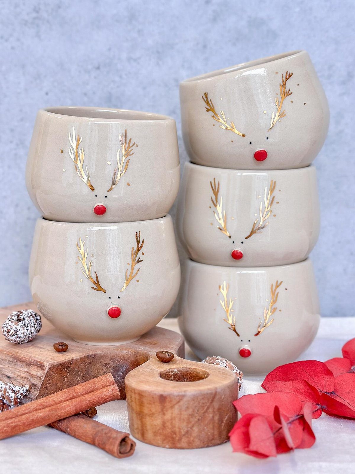 Beige ceramic mugs with gold reindeer antlers and red noses on wooden blocks.