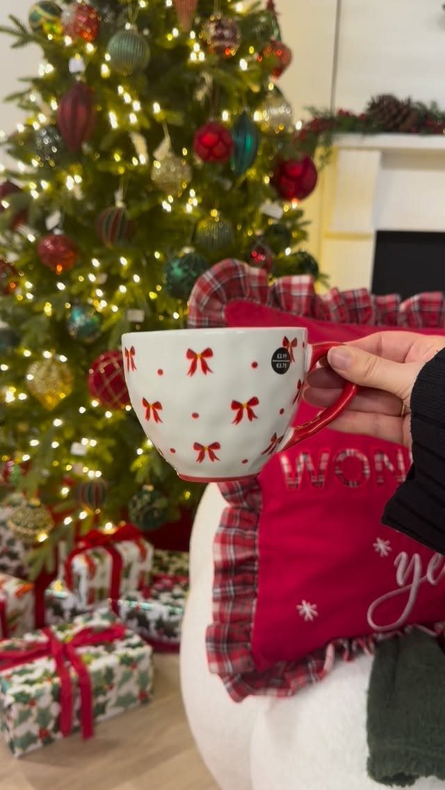 White coffee mug with red bows held in front of a decorated Christmas tree.