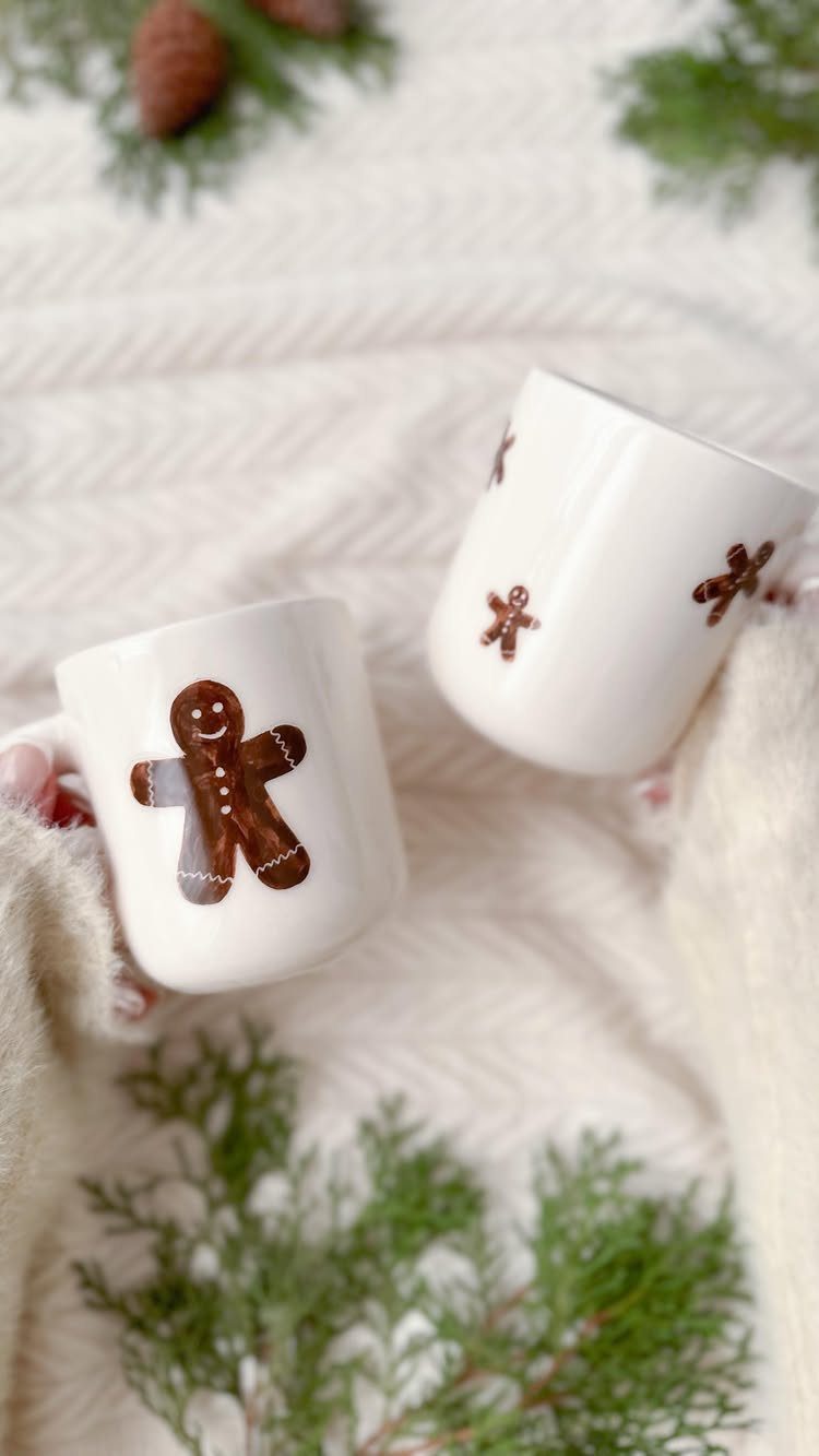 White ceramic mugs with hand-drawn gingerbread men on a white textured blanket.