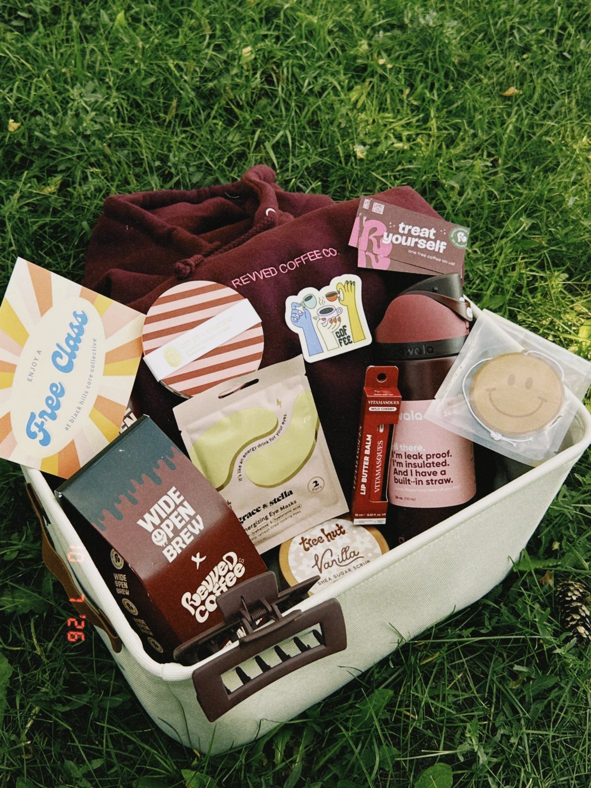 White gift basket on grass with coffee products, a maroon hoodie, and treats.