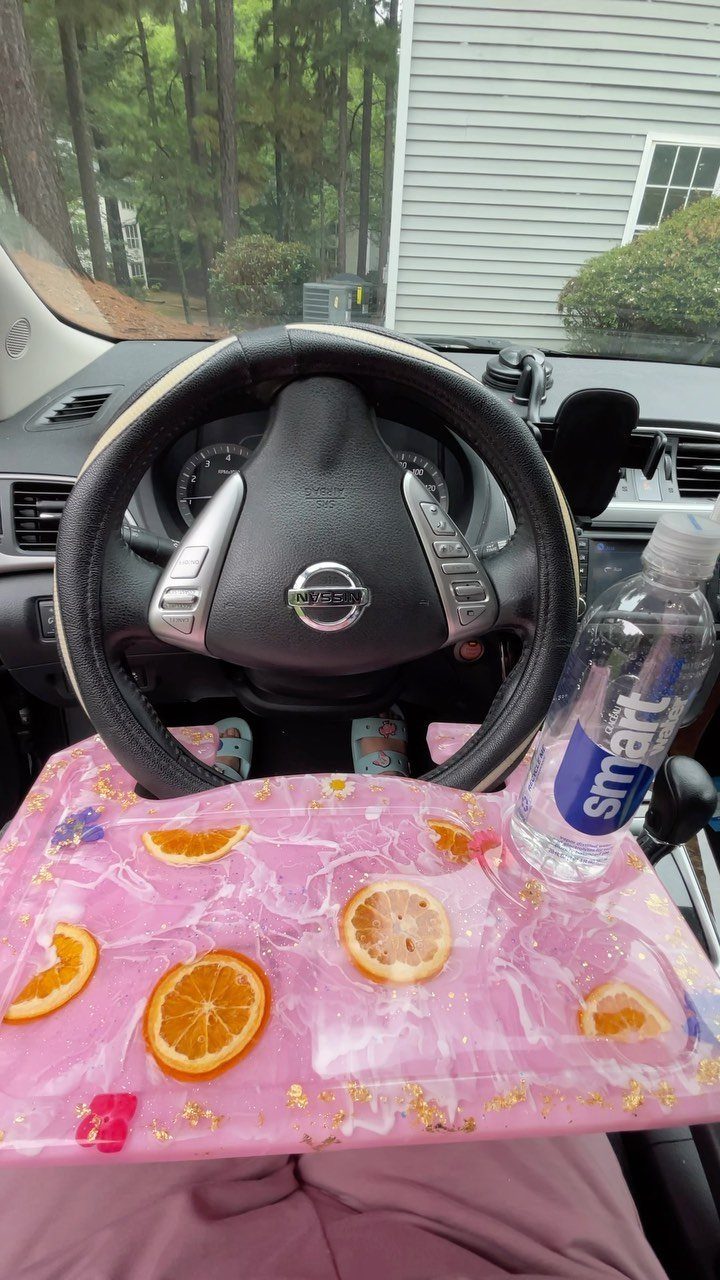 Pink resin steering wheel tray with orange slices and a water bottle inside a car