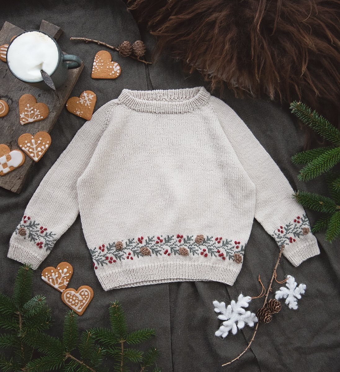 Cream knit sweater with festive berry embroidery surrounded by cookies and pine branches.