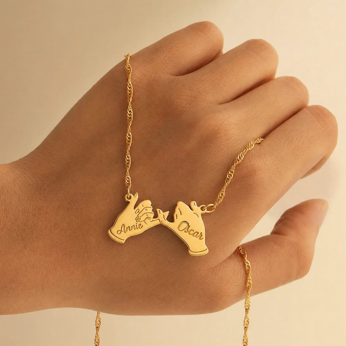 Gold pinky-swear necklace with names Annie and Oscar engraved, held in a hand.