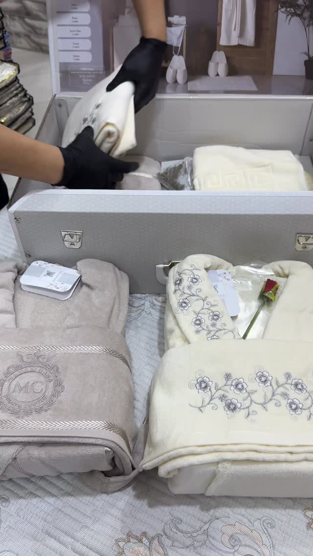 Embroidered white and beige blankets arranged in a grey box with a red rose.