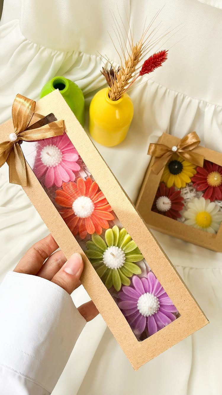 Flower-shaped colorful candles in brown gift boxes held over a white fabric background.