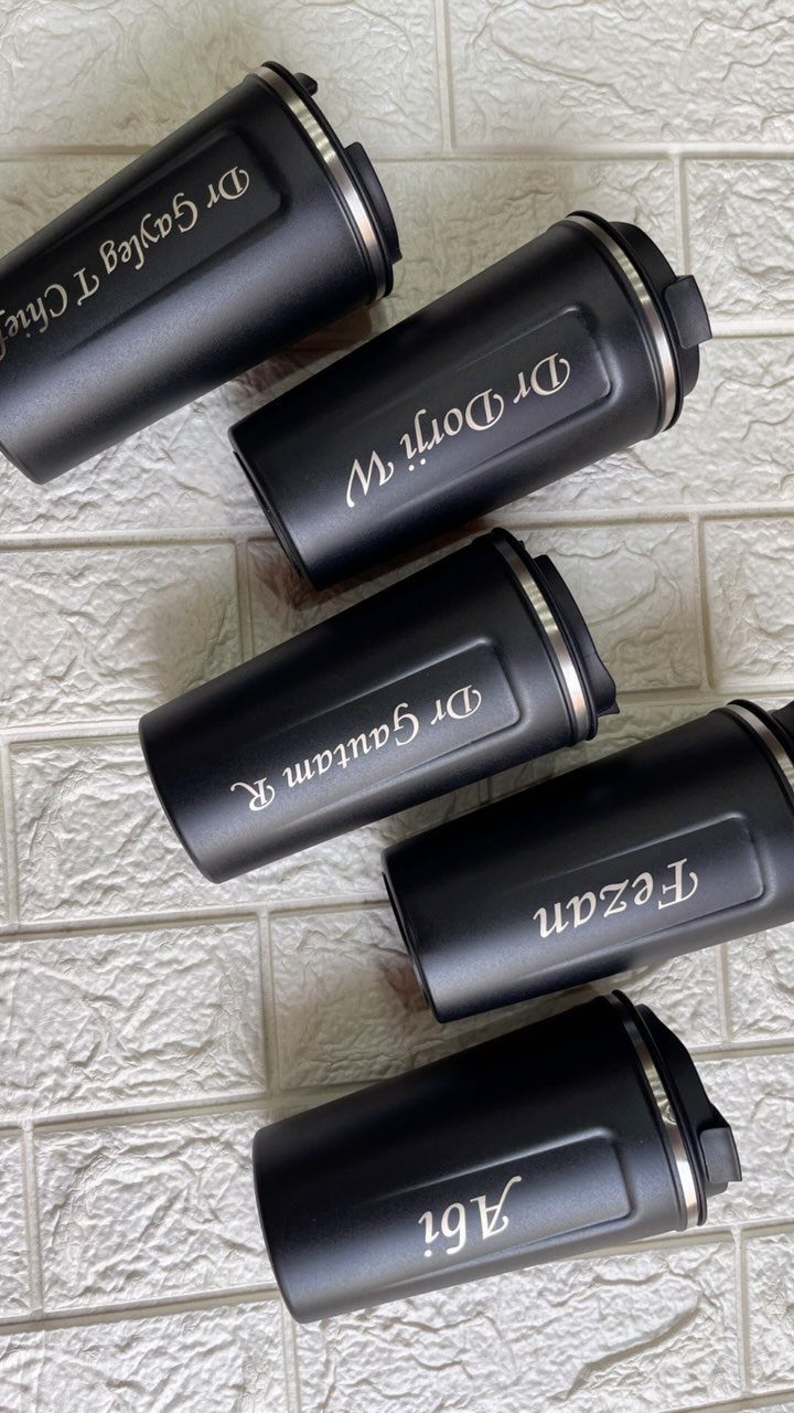 Five matte black insulated travel mugs with personalized names on a white brick background