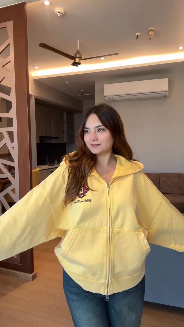 Woman wearing an oversized yellow zip-up hoodie and blue jeans in a living room.
