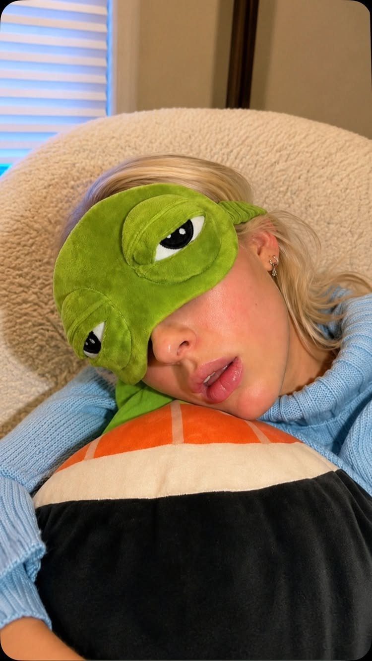 Woman wearing a green dinosaur sleep mask resting on a sushi plush pillow