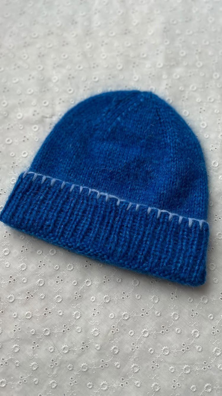 Blue knitted beanie hat resting on a white embroidered fabric background.