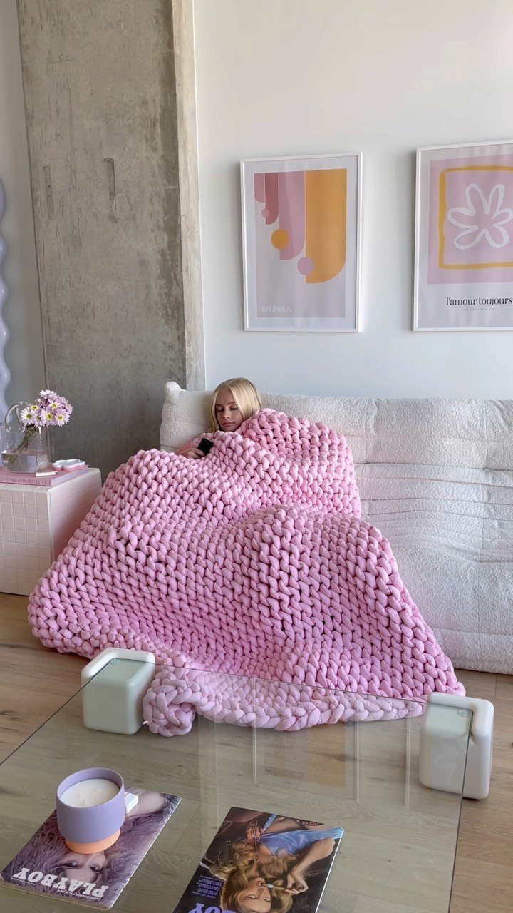 Woman wrapped in a chunky pink knit blanket sitting on a white sofa.