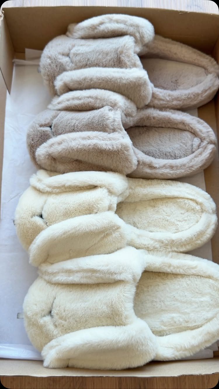 Four pairs of fuzzy cream and beige slippers inside a cardboard shipping box.