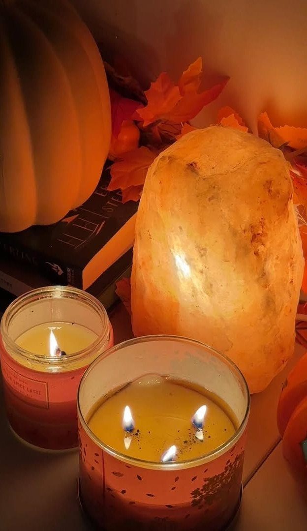Lit scented candles and a glowing salt lamp with autumn leaves and pumpkin.