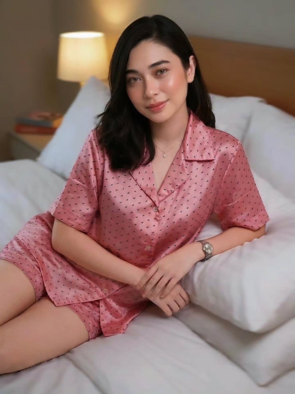 Woman wearing pink satin pajamas sitting on a white bed with warm lighting.