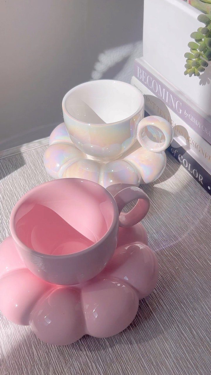 Pink and iridescent white ceramic mugs resting on cloud-shaped saucers next to books.