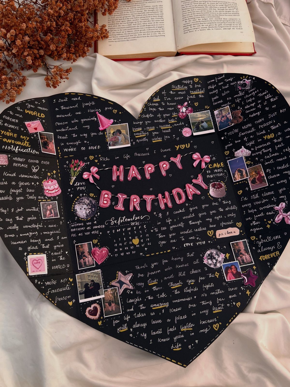 Heart-shaped black birthday board with photos, handwritten messages, and pink decorations on white fabric.