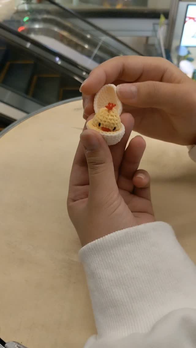 Tiny yellow crocheted chick in a white eggshell held by two hands.
