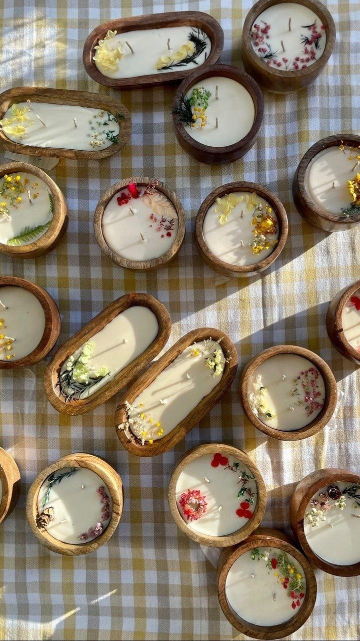 Handmade white candles in wooden bowls decorated with dried flowers on checkered fabric