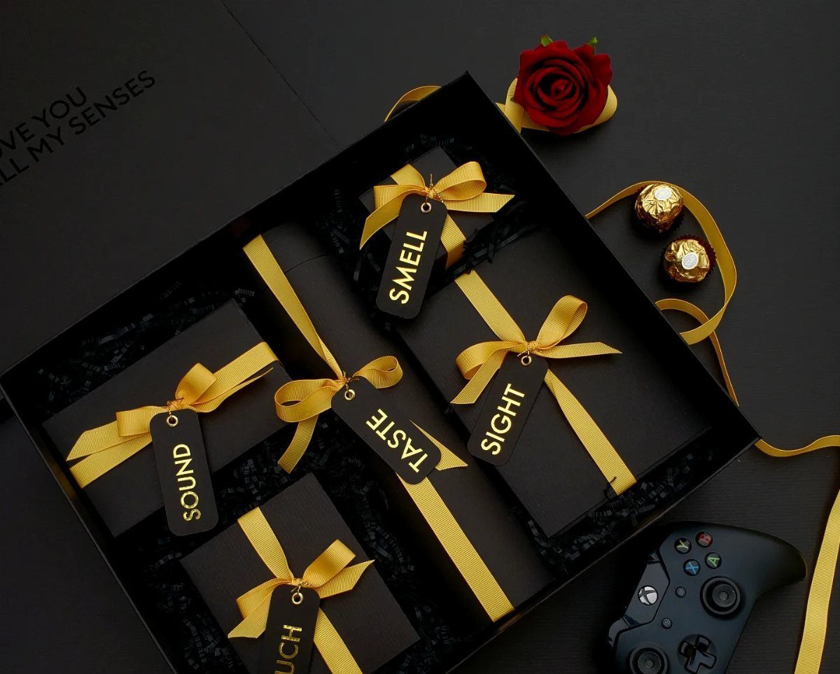 Five black gift boxes labeled with the five senses, accompanied by a rose and chocolates.