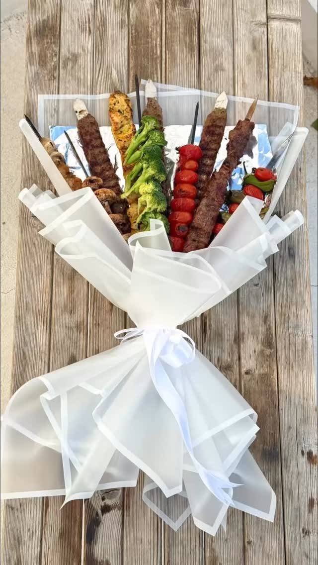 Grilled meat and vegetable skewers arranged as a bouquet wrapped in white fabric.