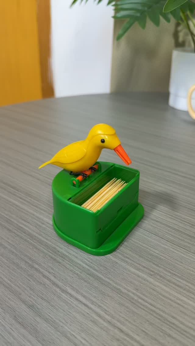 Yellow plastic bird perched on a green toothpick holder on a grey table