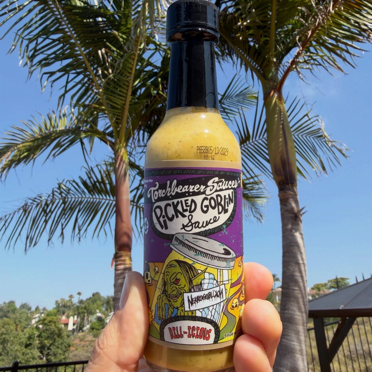 Bottle of Pickled Goblin hot sauce held against a background of palm trees.