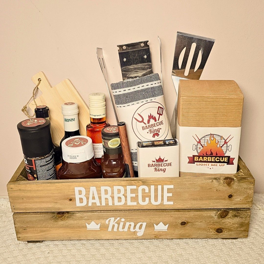 Wooden barbecue gift crate containing grilling tools, sauces, and spices on a white surface.