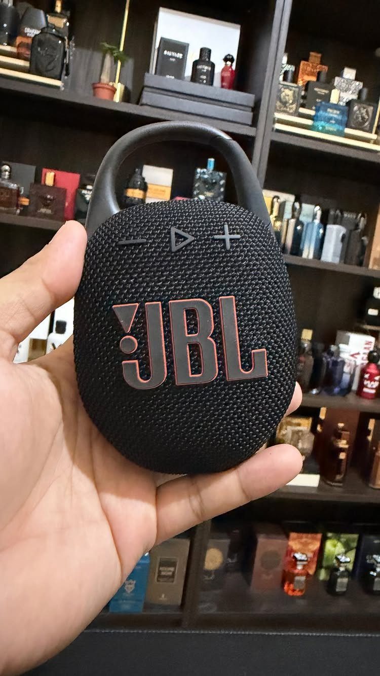 Black JBL portable speaker held in a hand against a background of perfumes.