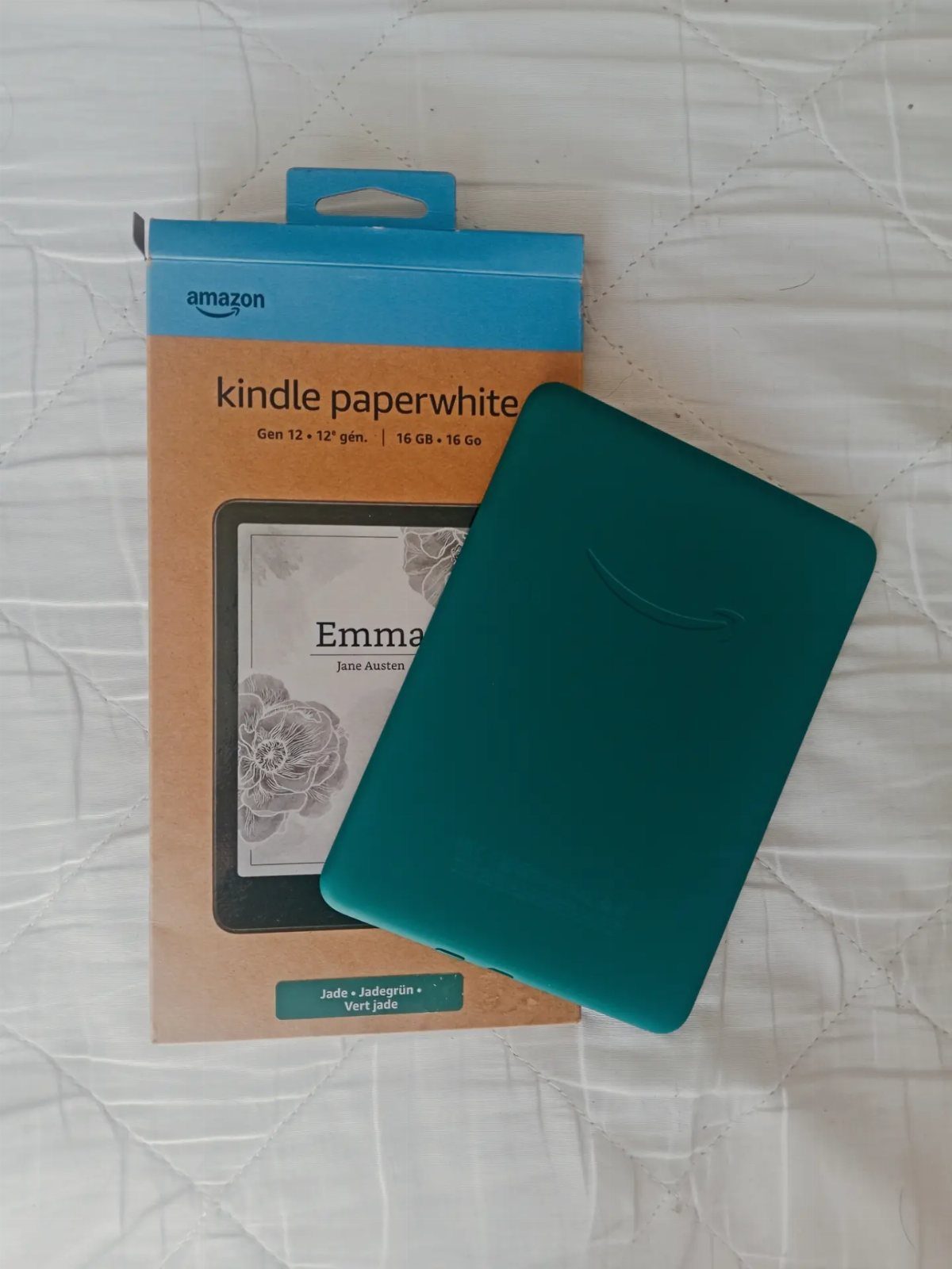 Teal Kindle Paperwhite e-reader and its packaging on a white quilted background.