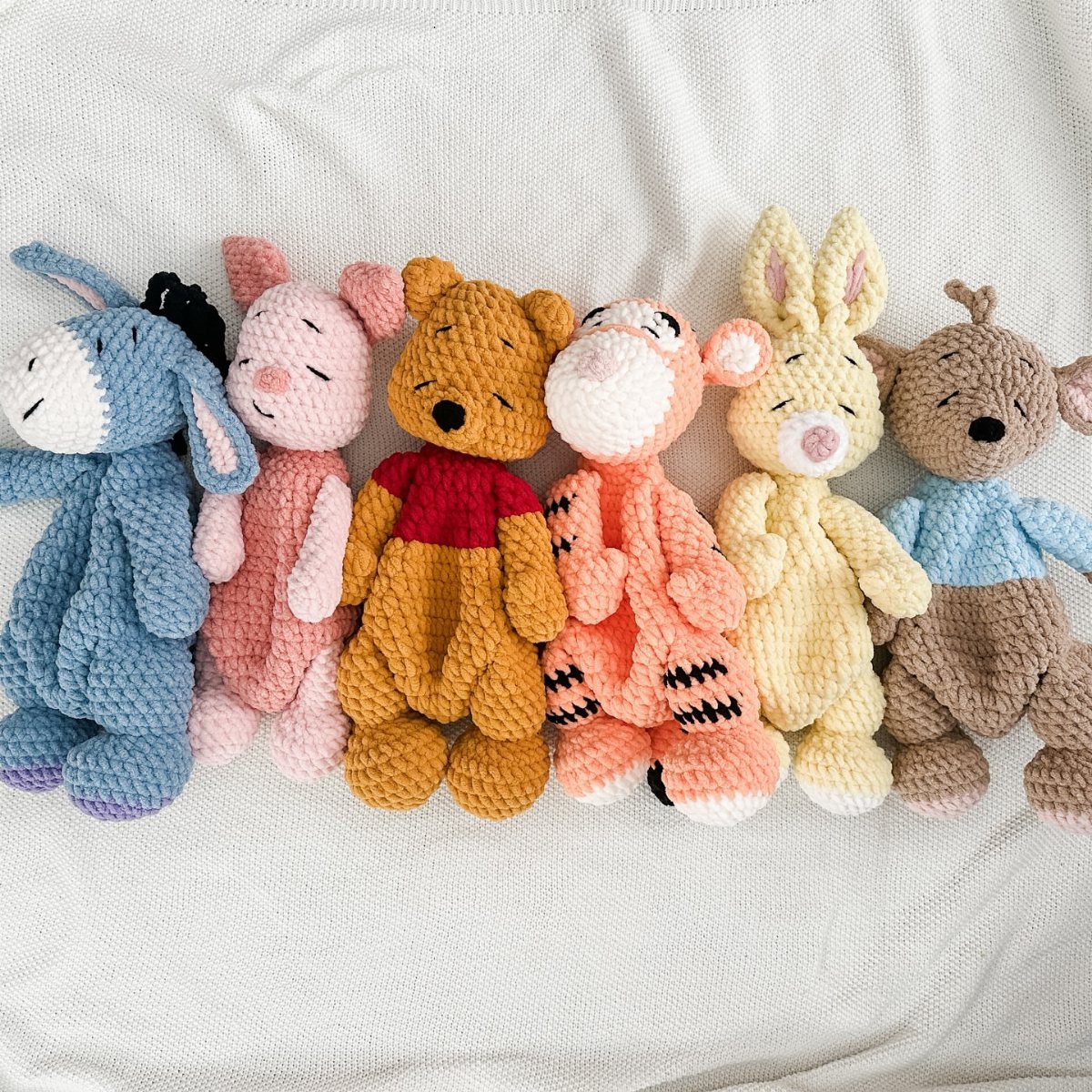 Six crocheted Winnie the Pooh characters sitting in a row on white fabric.