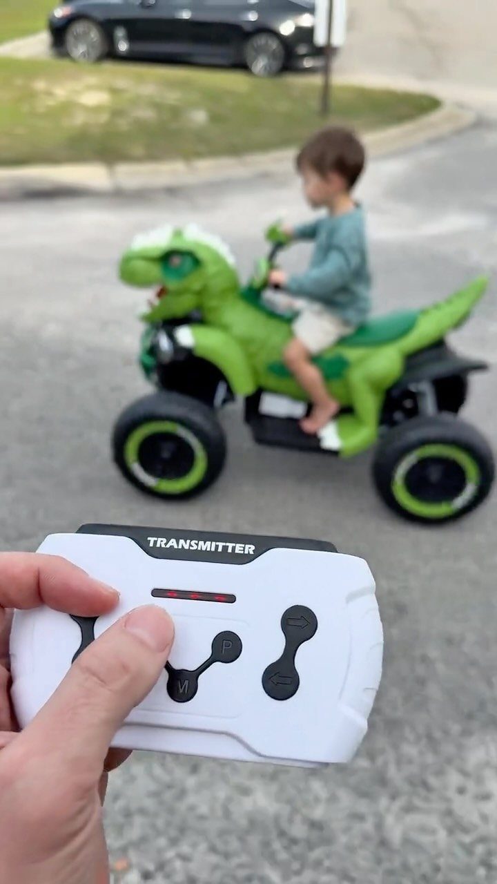 Child riding a green dinosaur ride-on toy controlled by a white remote transmitter.