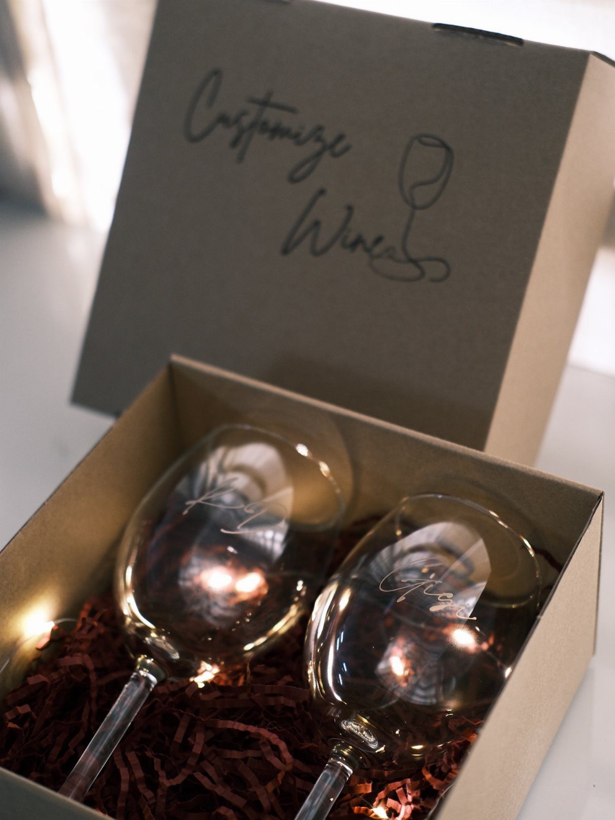 Two engraved wine glasses resting on red filler inside a brown gift box.
