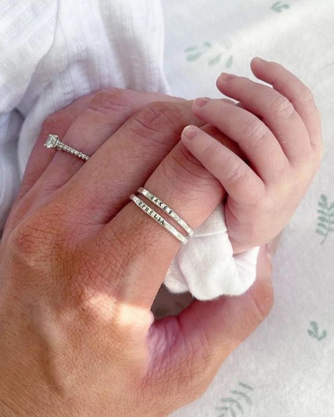 Adult hand holding a baby's hand wearing silver personalized stacking name rings.