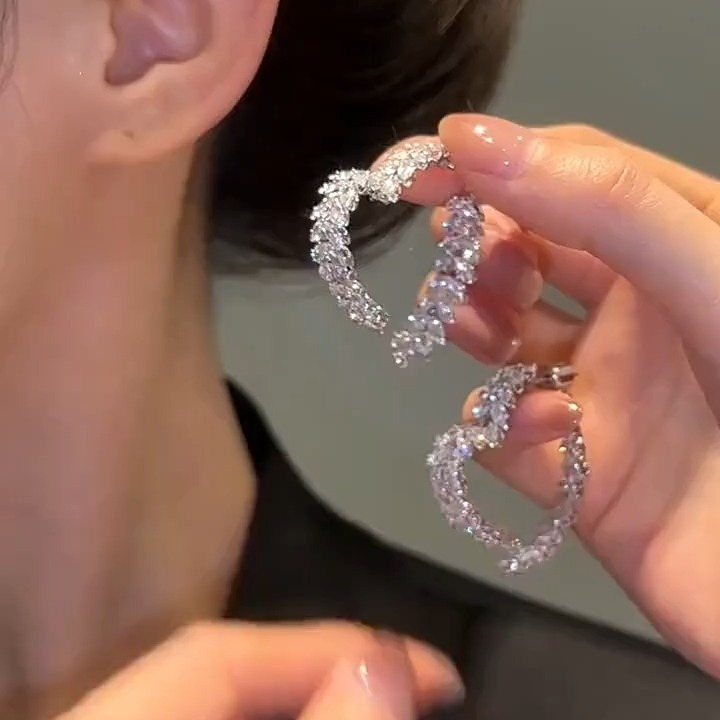 Heart-shaped silver hoop earrings with sparkling crystals held by a person's hand.