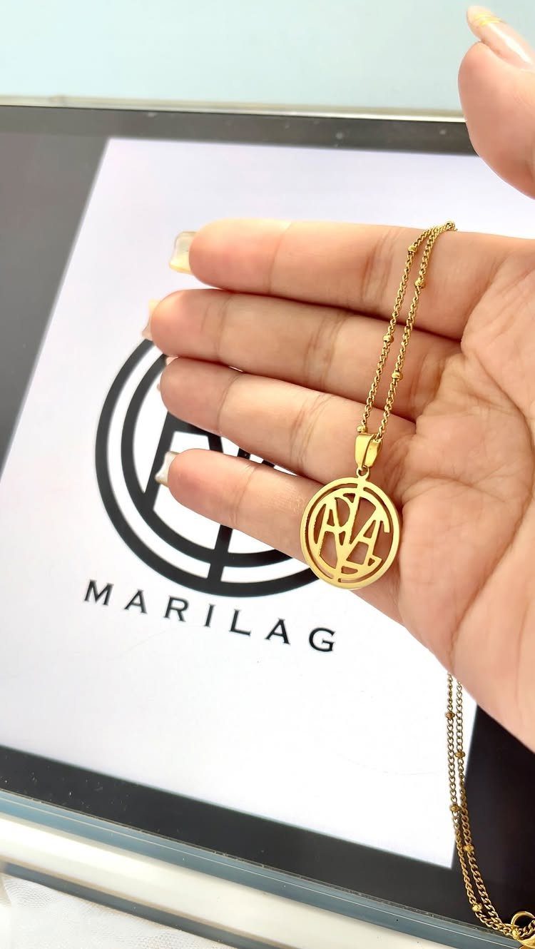 Gold monogram pendant necklace held in a hand over a branded logo background.