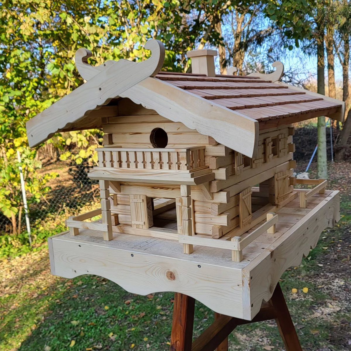 Ornate wooden birdhouse shaped like a log cabin on a wooden stand outdoors.