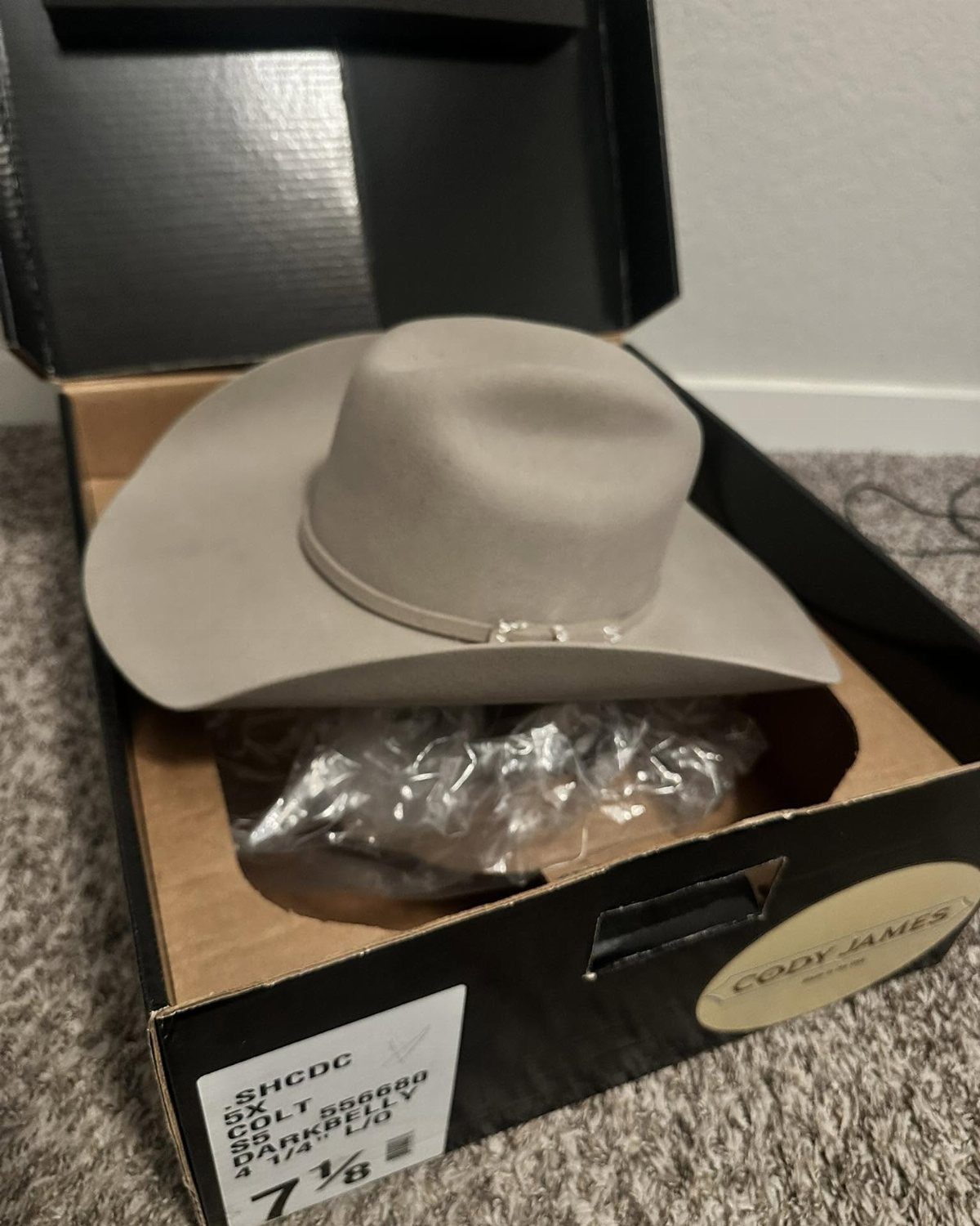 Light grey cowboy hat inside a black Cody James branded box on carpet.