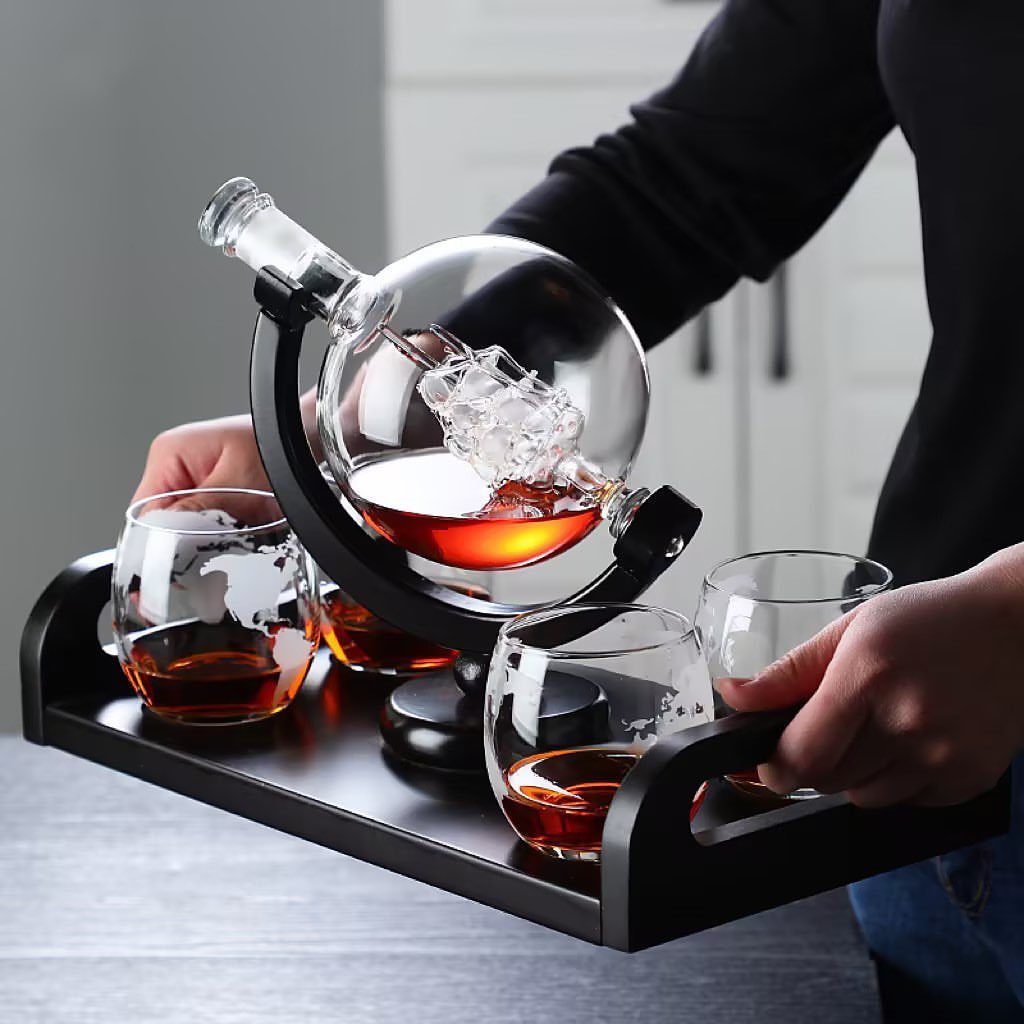 Globe-shaped whiskey decanter and matching glasses on a black tray held by hands.