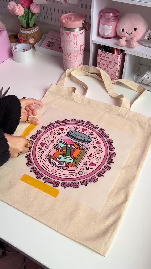 Hands applying a book-themed transfer design to a beige canvas tote bag.