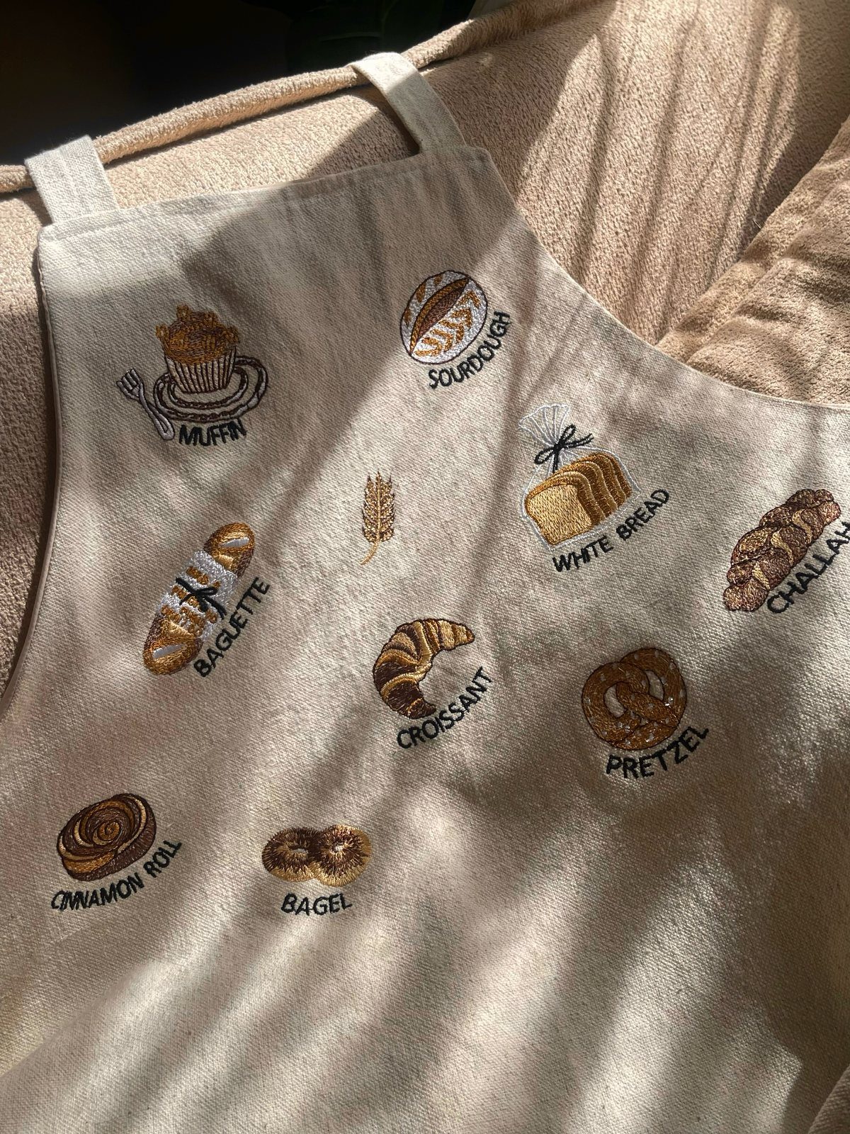 Beige linen apron with embroidered bakery items like croissants, baguettes, and pretzels.