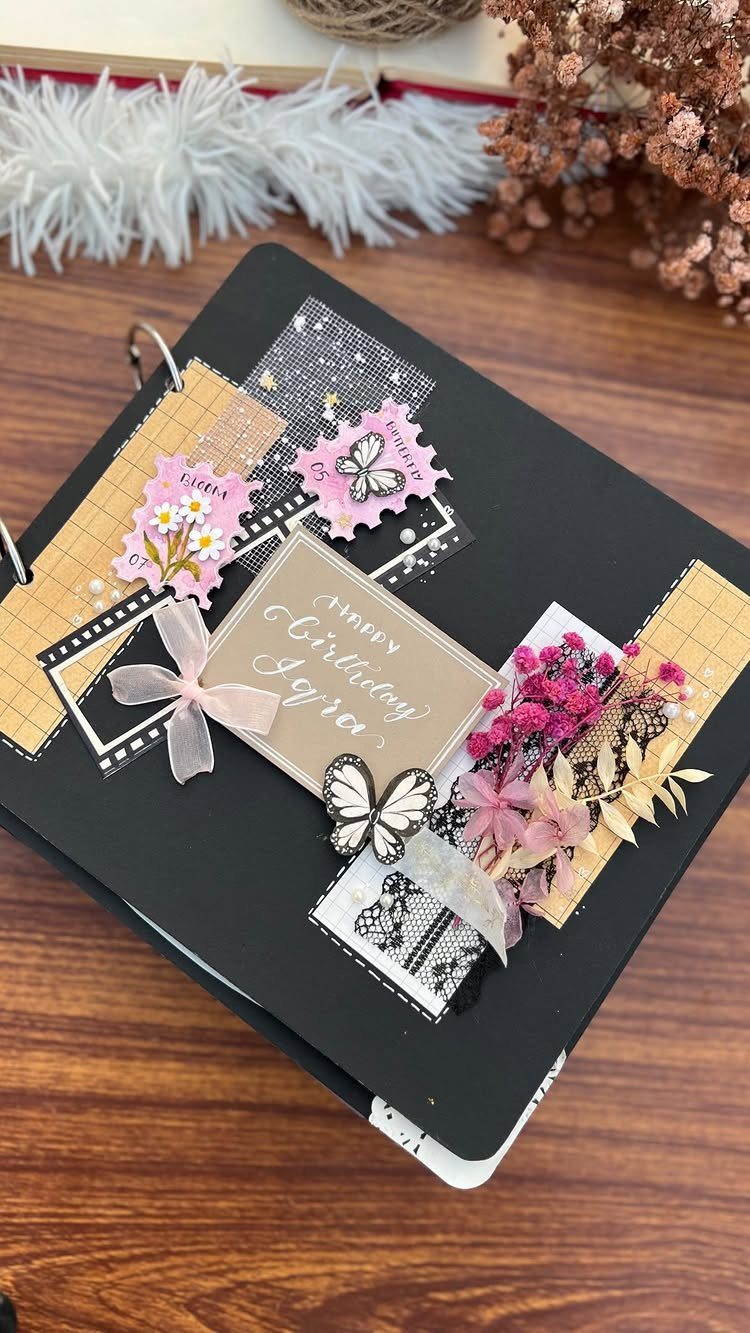 Decorated black sketchbook with birthday card, pink flowers, and butterfly on wooden table