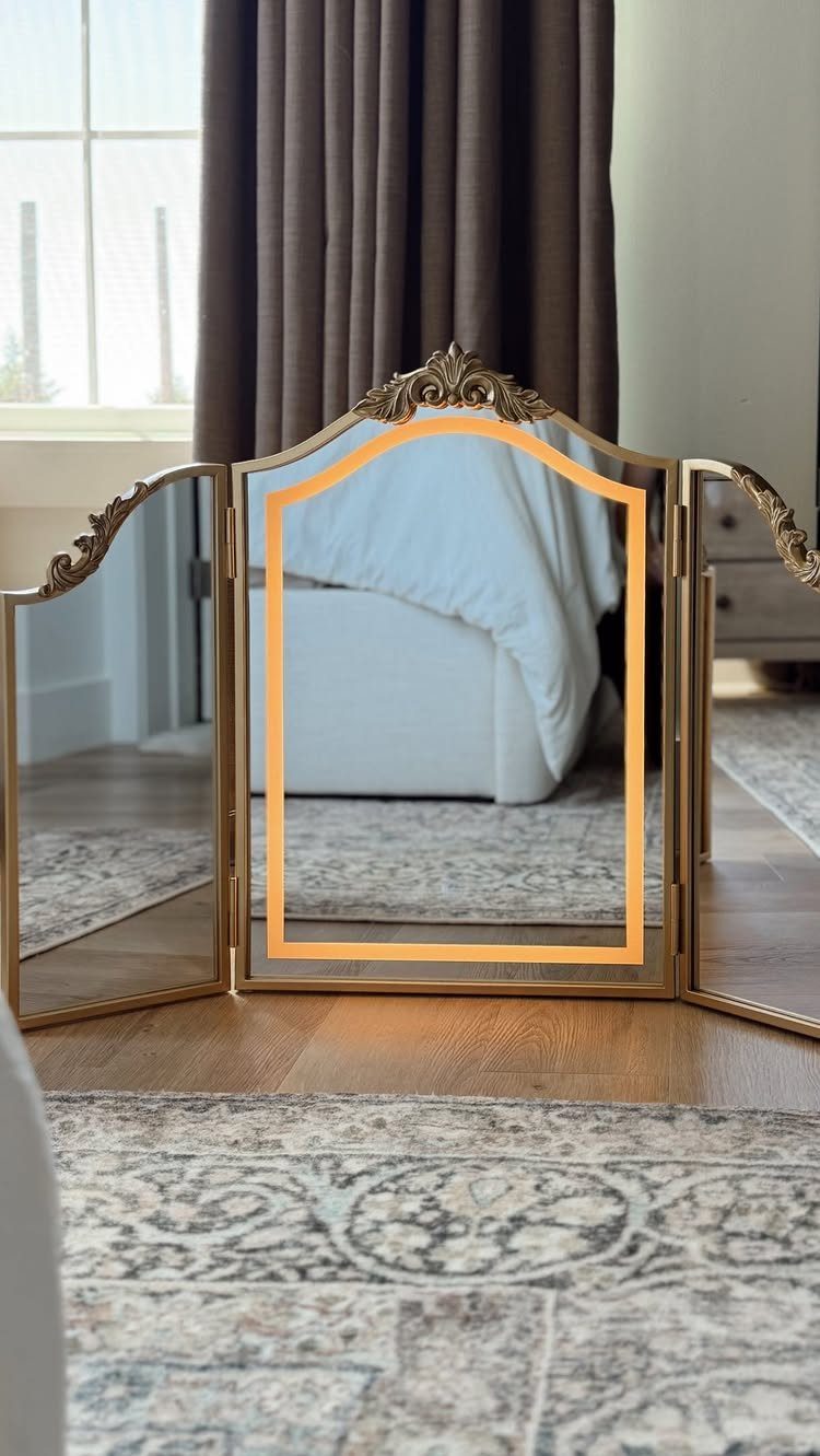 Gold tri-fold vanity mirror reflecting a bedroom with a white bed and curtains.