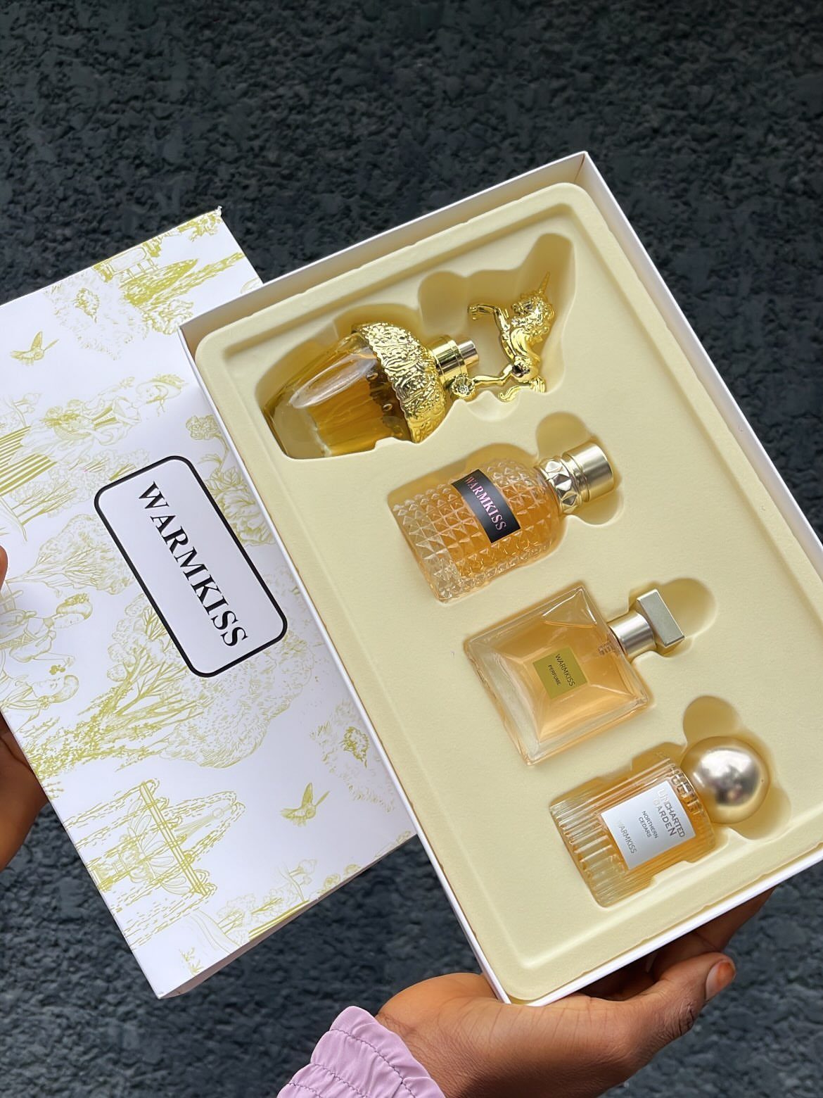 Four gold-toned perfume bottles in a cream gift box held by a person